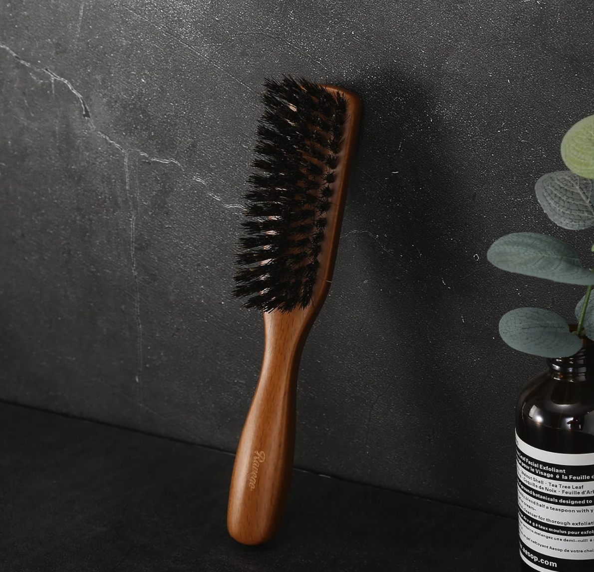 Ravego Boar Bristle Beard Brush on a dark surface with a plant in the corner