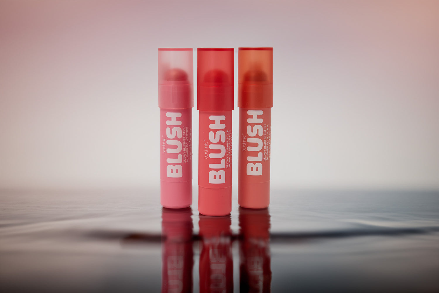 Three tubes of BLUSH from Technic Cosmetics in pink, red, and orange shades, standing upright on a reflective dark surface against a soft pink gradient background.