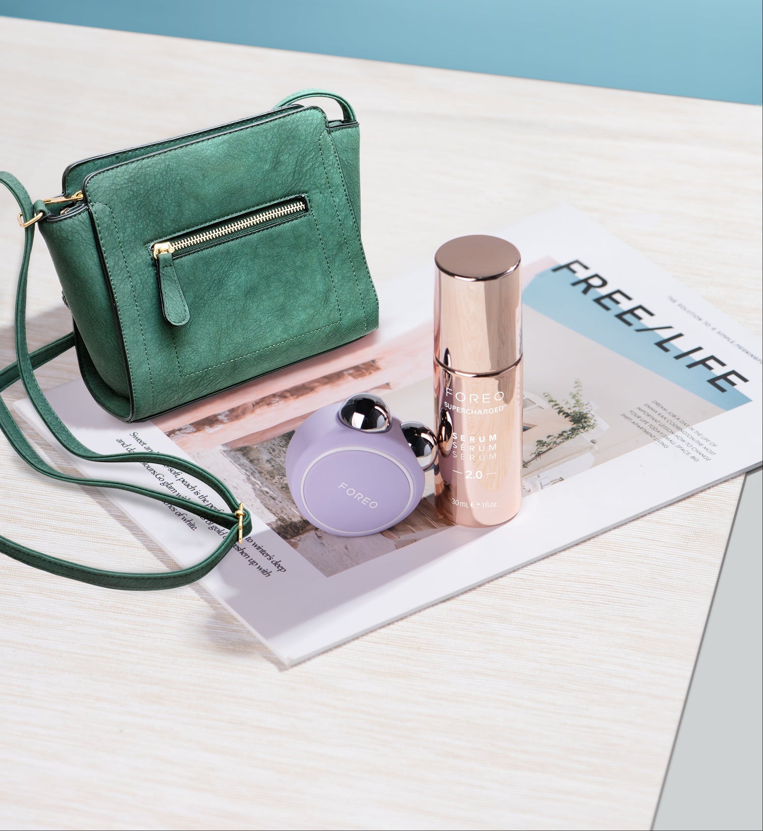 A green suede crossbody handbag with gold hardware and a matching strap, styled on a magazine next to a pink cylindrical cosmetic bottle and a small lavender foreo bear 2 go skin tool device/