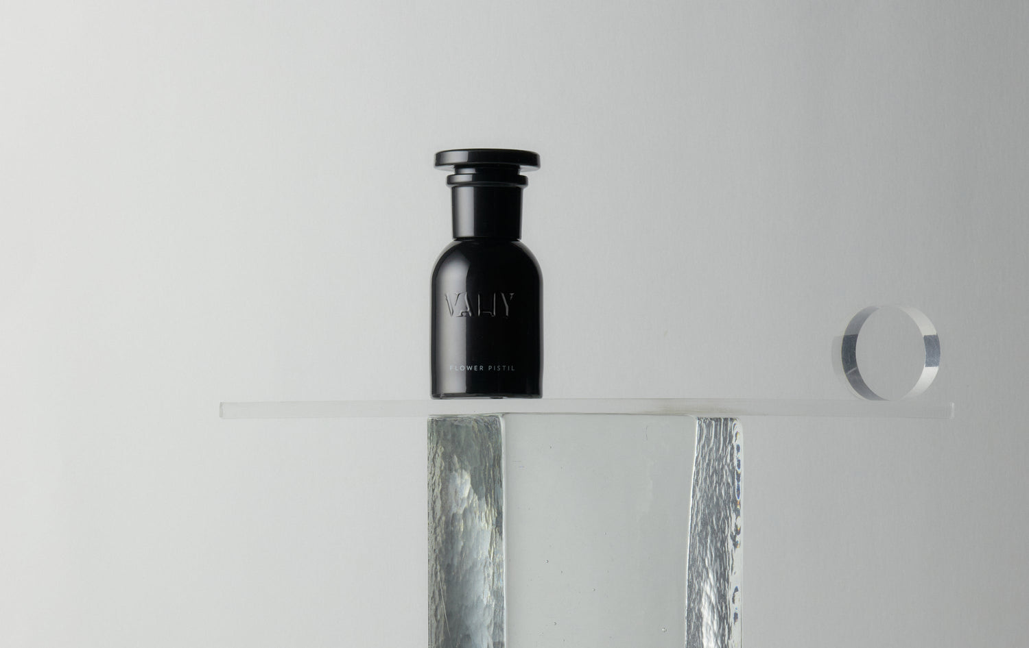 A minimalist studio shot of a 50ml black glass bottle of perfume with a black cap, featuring the brand name 'Vahy on the front, displayed on a clear acrylic shelf with a textured silver column and mirror reflection. Scent Flower Pistil.