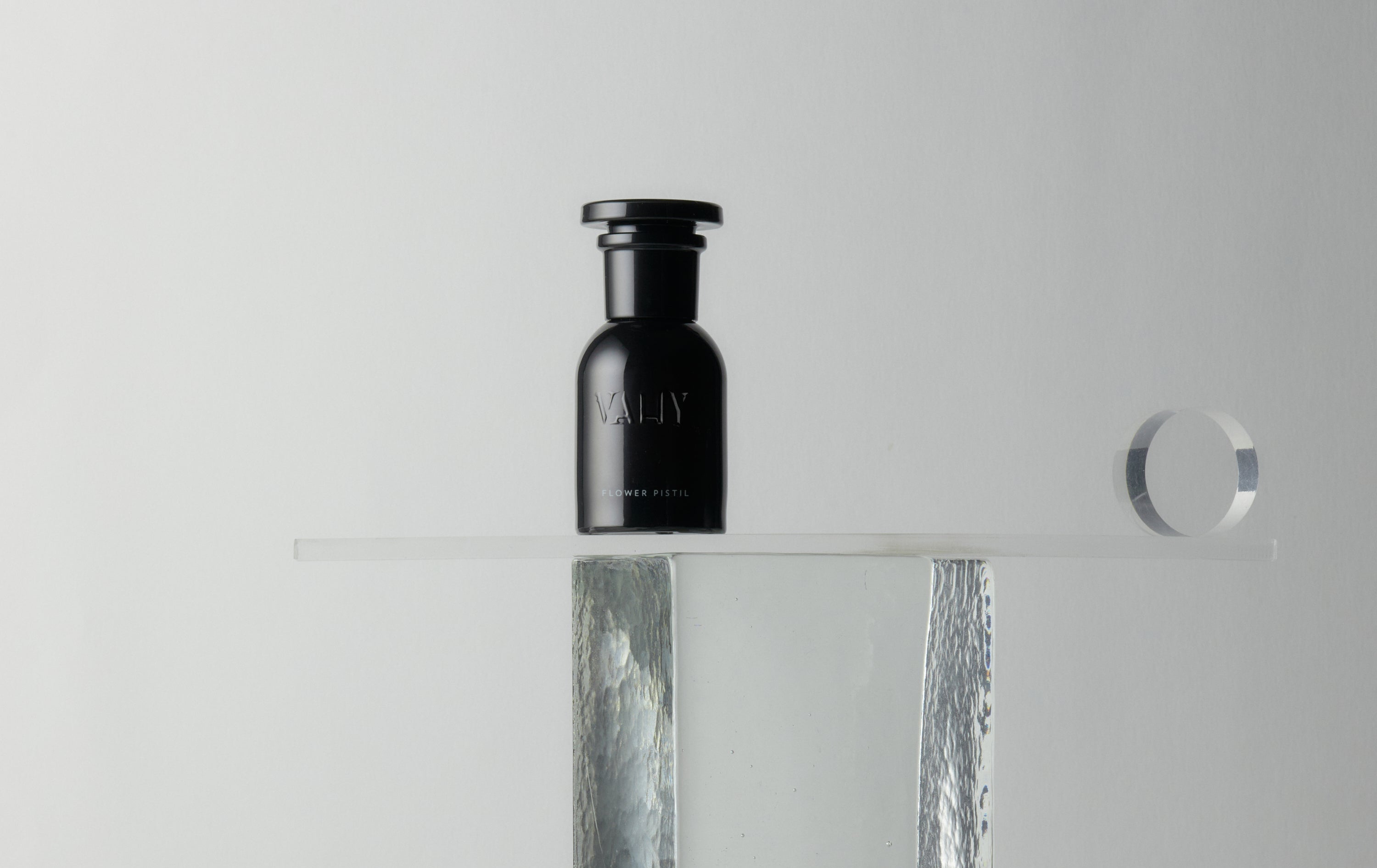 A minimalist studio shot of a 50ml black glass bottle of perfume with a black cap, featuring the brand name 'Vahy on the front, displayed on a clear acrylic shelf with a textured silver column and mirror reflection. Scent Flower Pistil.