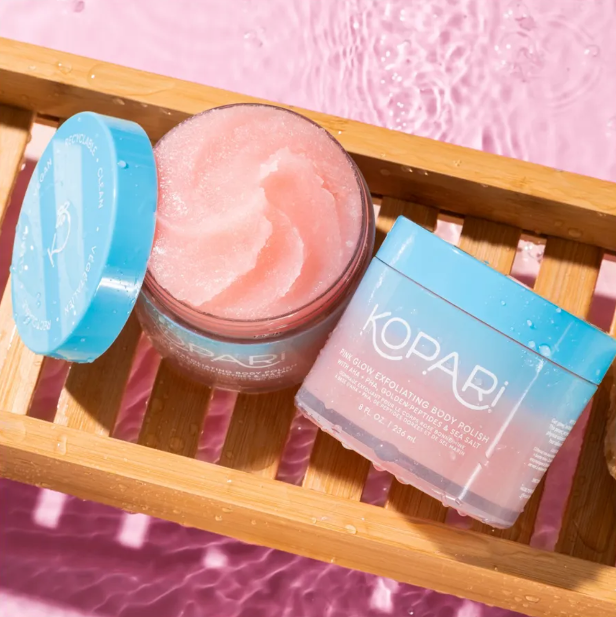 Kopari body scrub jar with pink scrub and blue lid on a wooden tray against a pink watercolor background