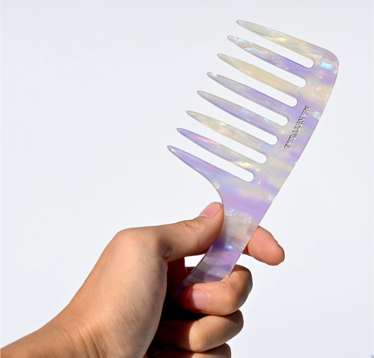 Hand holding manetural iridescent  wide tooth comb against a light gray background
