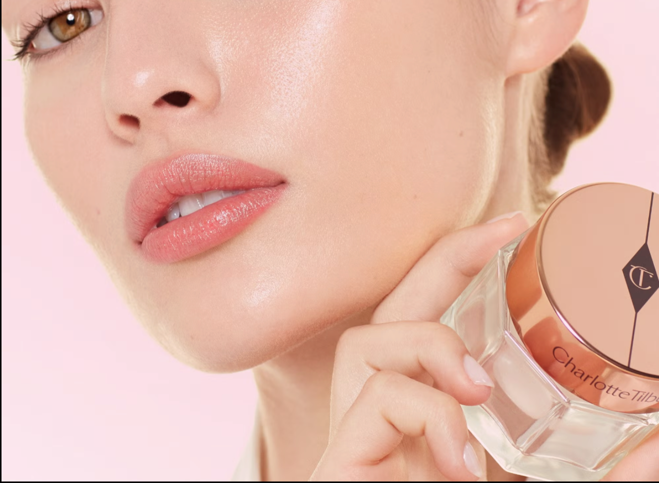 Close-up of a white woman holding the charlotte tilbury magic cream to her face on a pink background.