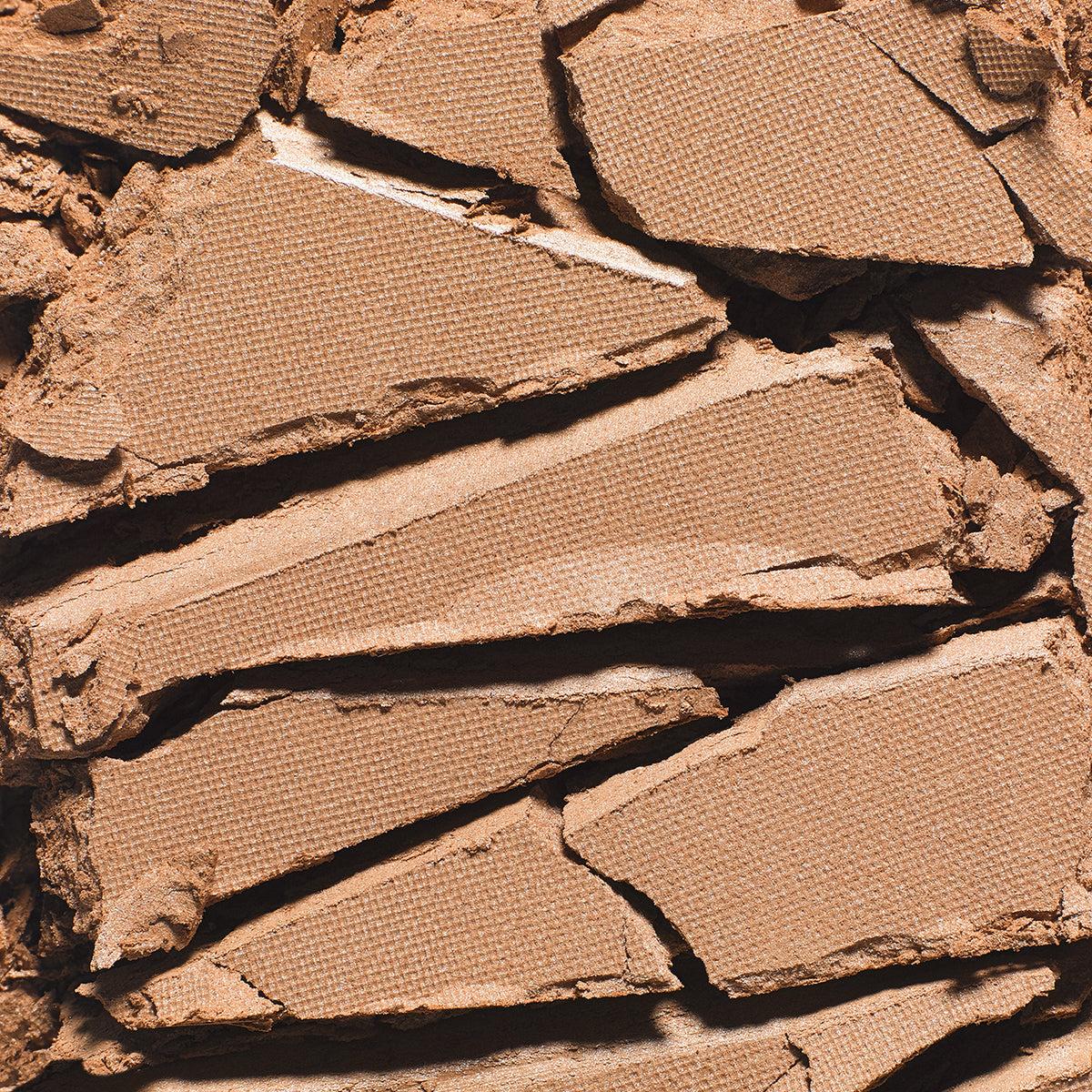 A close-up of a cracked, broken pressed powder makeup pan showing the textured brown pigment and fine powder makeup contour.